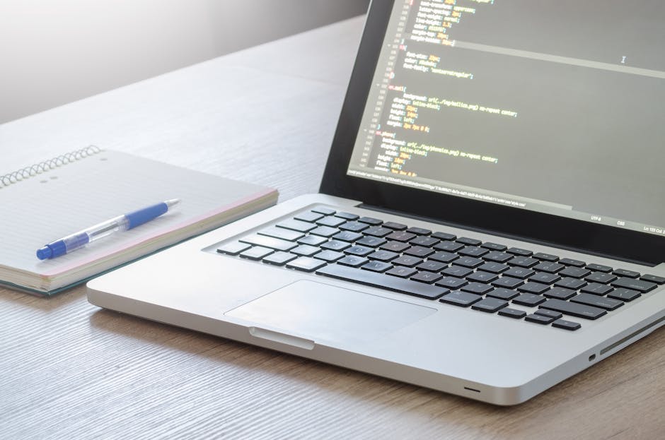 A close-up view of a modern silver laptop with a black keyboard displaying lines of colorful programming code on a screen, placed on a light wooden desk. Next to the laptop is an open spiral-bound not
