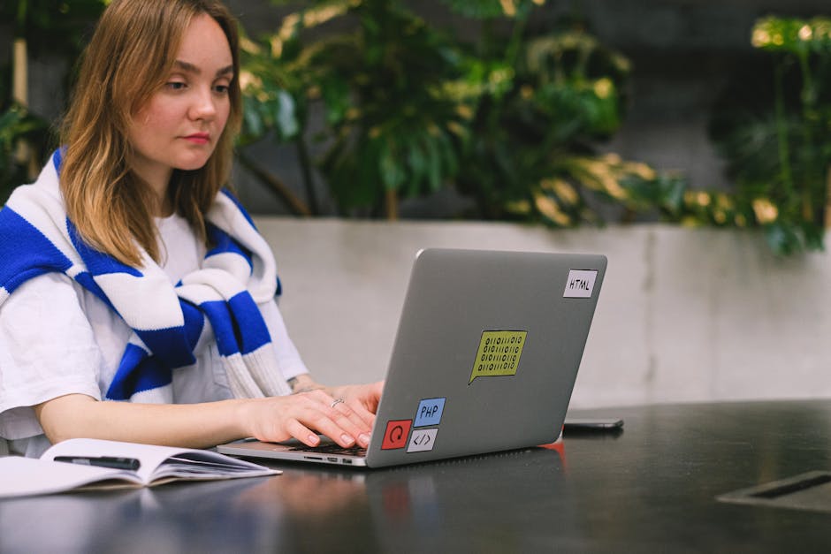 A young woman with shoulder-length light brown hair, dressed in a white shirt and a blue and white striped scarf draped over her shoulders, seated outdoors at a dark table with a laptop in front of he